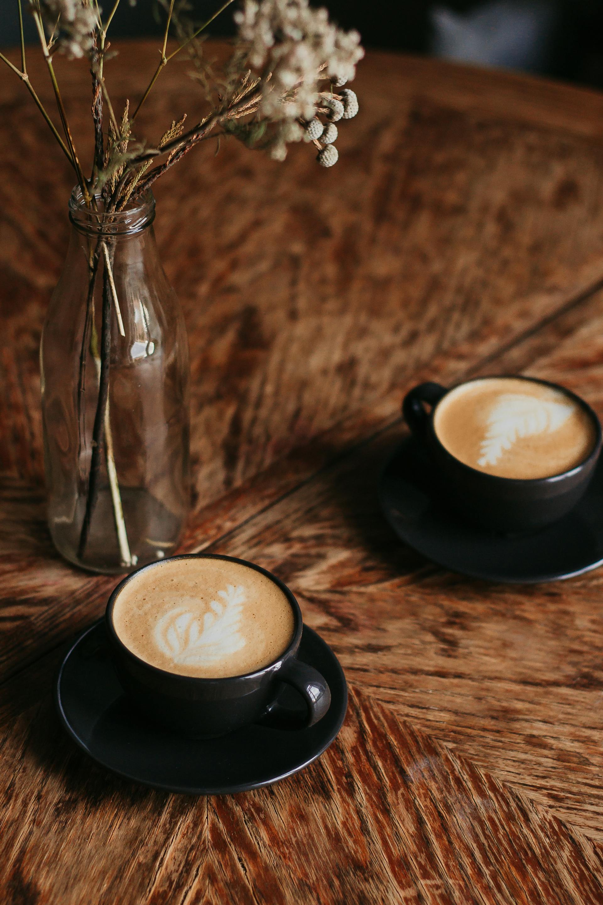 Two cappuccinos on a wooden table beside a small jar of dried flowers
