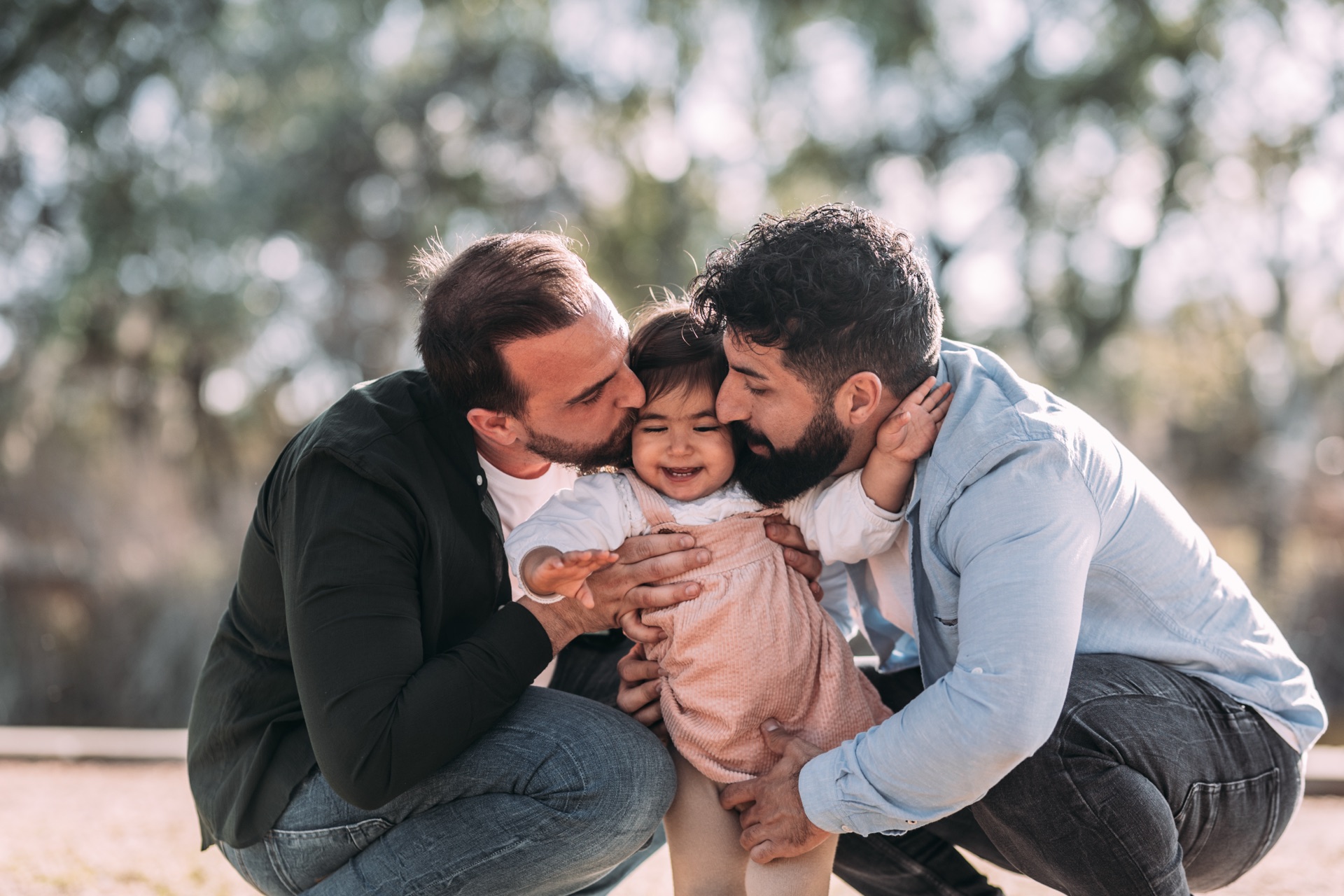 Two men tenderly holding and kissing their smiling baby daughter in a sunlit park